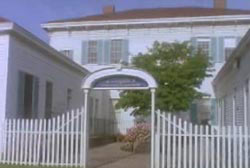 Drum barracks. Three white building surroundings a court yard with a tree in the center behind a white picket fence.